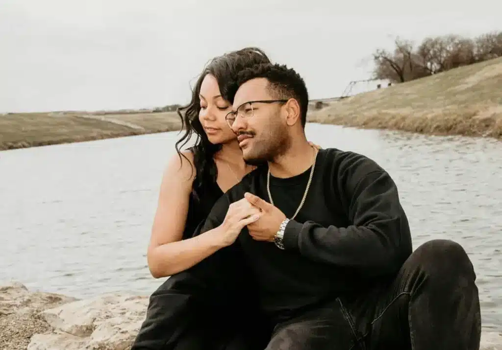 A young adult man and woman sitting closely together beside the water.